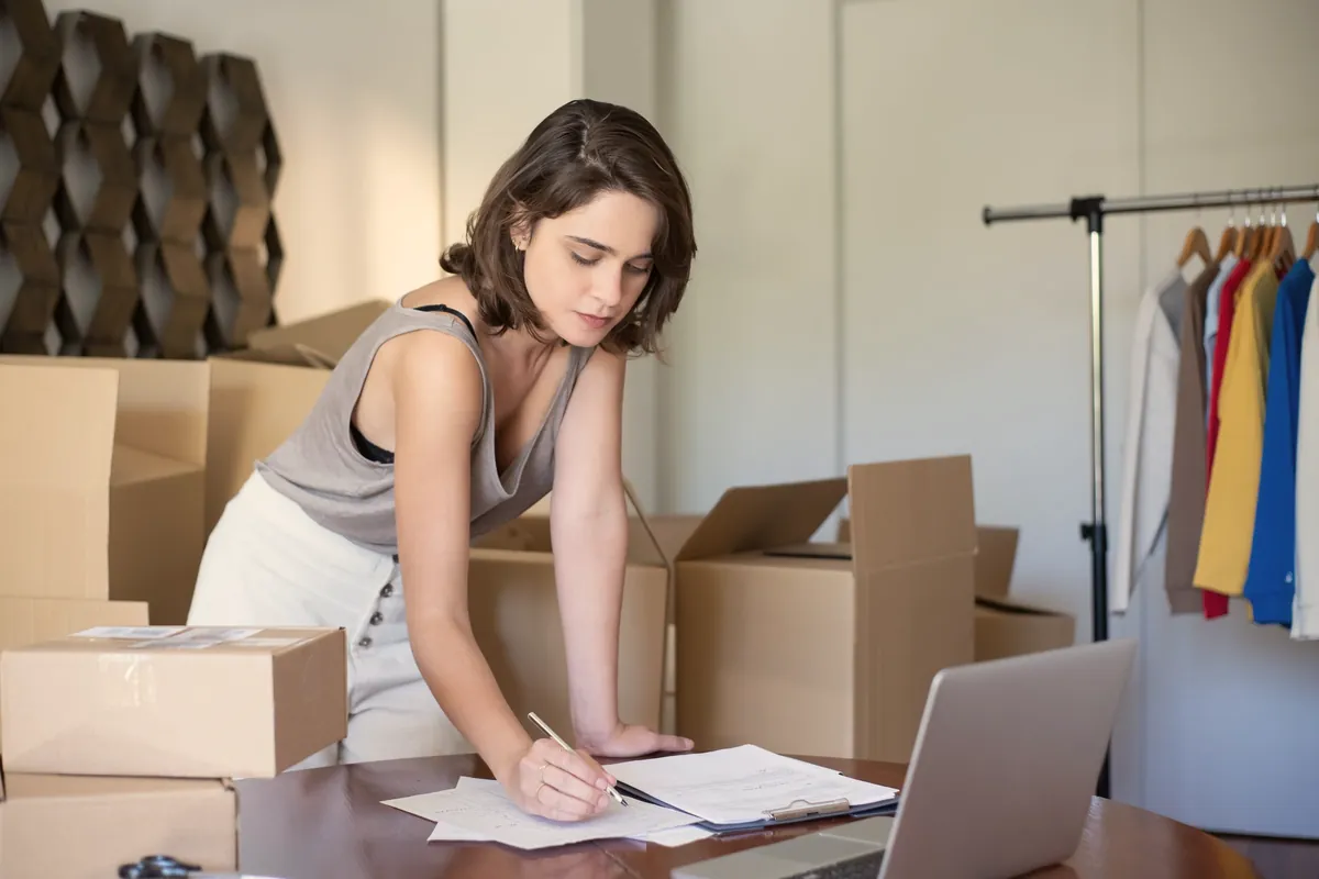 صاحب یک فروشگاه اینترنتی میان جعبههای سفارش، که کنار لپتاپ سفارشها را یادداشت میکند